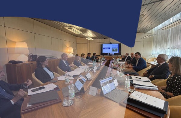 Business professionals sit around a long conference table in a modern meeting room, with laptops, documents, and water bottles as a presentation plays on a screen at the far end.