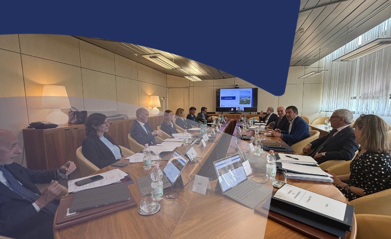 Business professionals sit around a long conference table in a modern meeting room, with laptops, documents, and water bottles as a presentation plays on a screen at the far end.