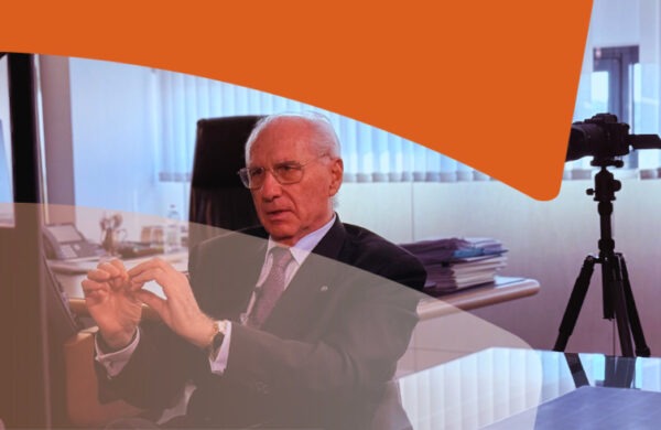 Older man in a suit explaining a point in an office, with a video camera on a tripod filming nearby.