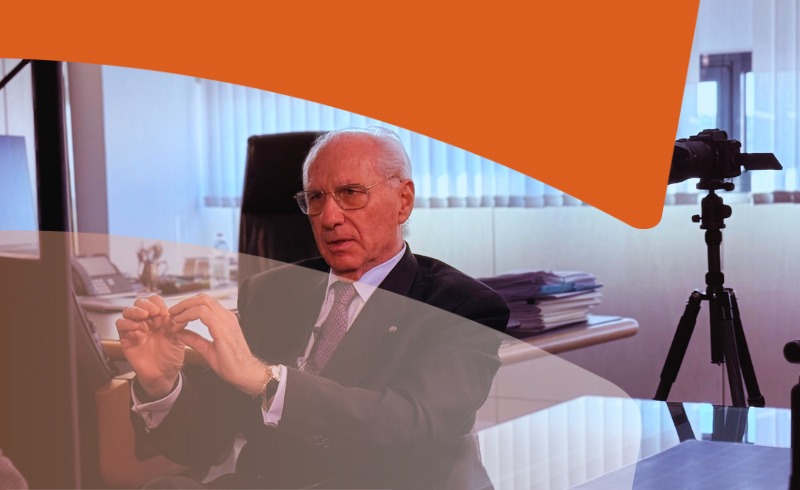 Older man in a suit explaining a point in an office, with a video camera on a tripod filming nearby.
